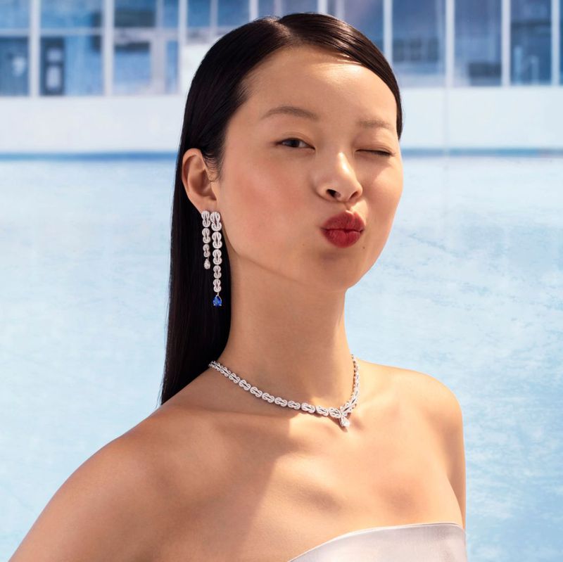 Close-up of a young Asian woman with dark hair, wearing elegant jewelry, winking, and puckering her lips, against a blue backdrop.