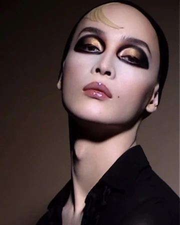 Striking beauty portrait: person with black head covering, dark eye makeup, light skin, and a black shirt against a brown backdrop.