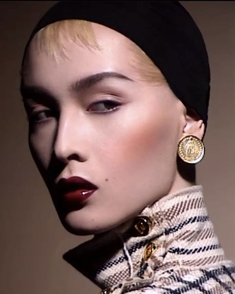 Close-up portrait of a blonde woman with dark lipstick and a black head covering wearing gold coin earrings and a collared jacket.