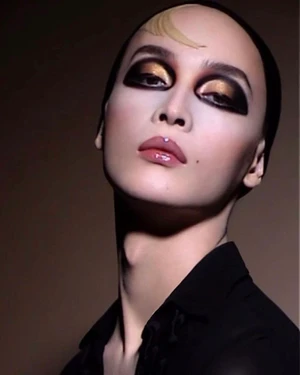 Close-up beauty portrait of a model with dramatic black and gold eye makeup, glossy lips, and a black shirt against a dark backdrop.