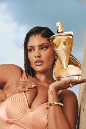Adult woman poses with gold perfume bottle in a studio shot against a blue sky background. The model wears peach-colored attire.