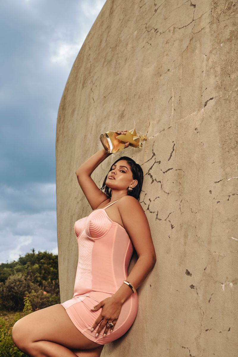 Brunette adult female in a peach-colored dress leans against a textured wall, holding a gold vase in an outdoor setting under a cloudy sky.