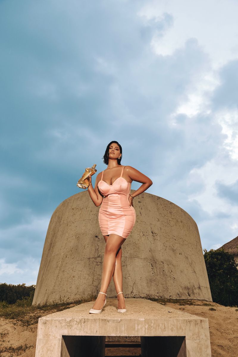 A woman poses in a peach dress and heels, holding a gold object, set against a backdrop of cloudy sky and a dome-shaped structure.