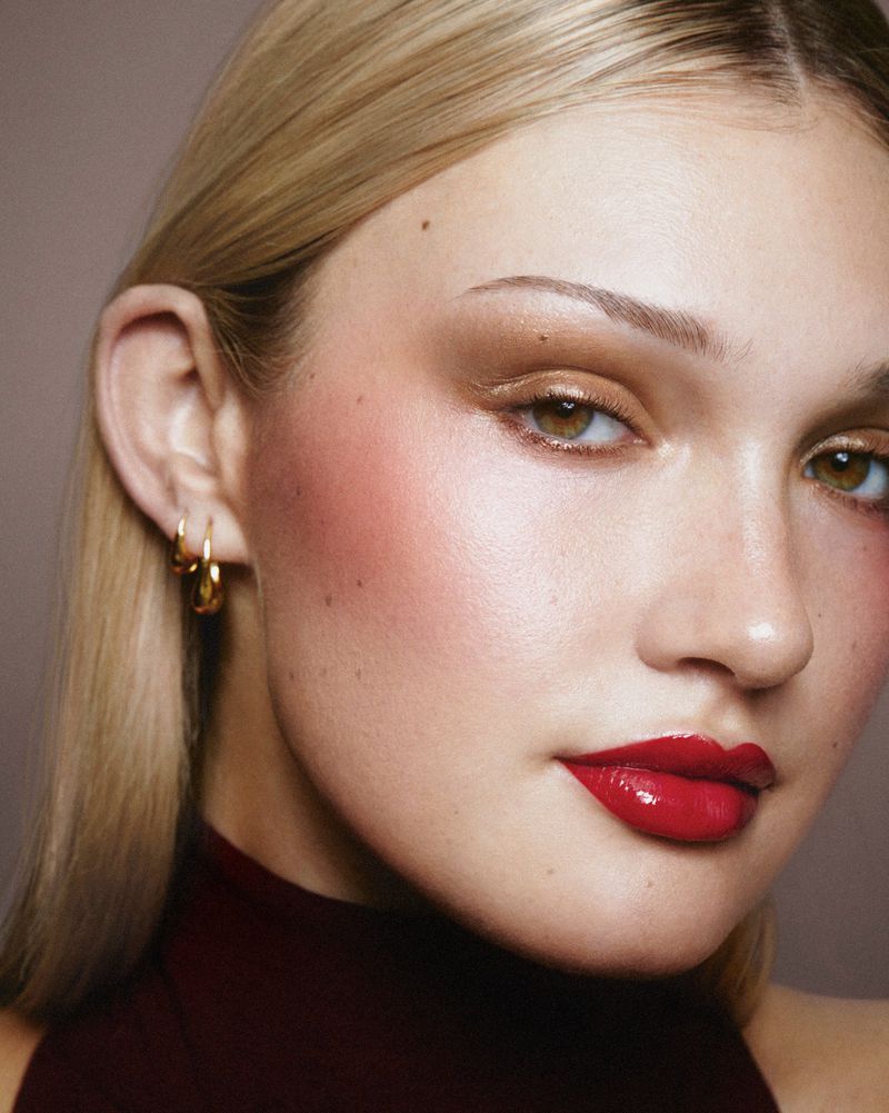 Close-up portrait of a blond woman with bold makeup including red lipstick, accentuated eyebrows, and blush, against a neutral background.