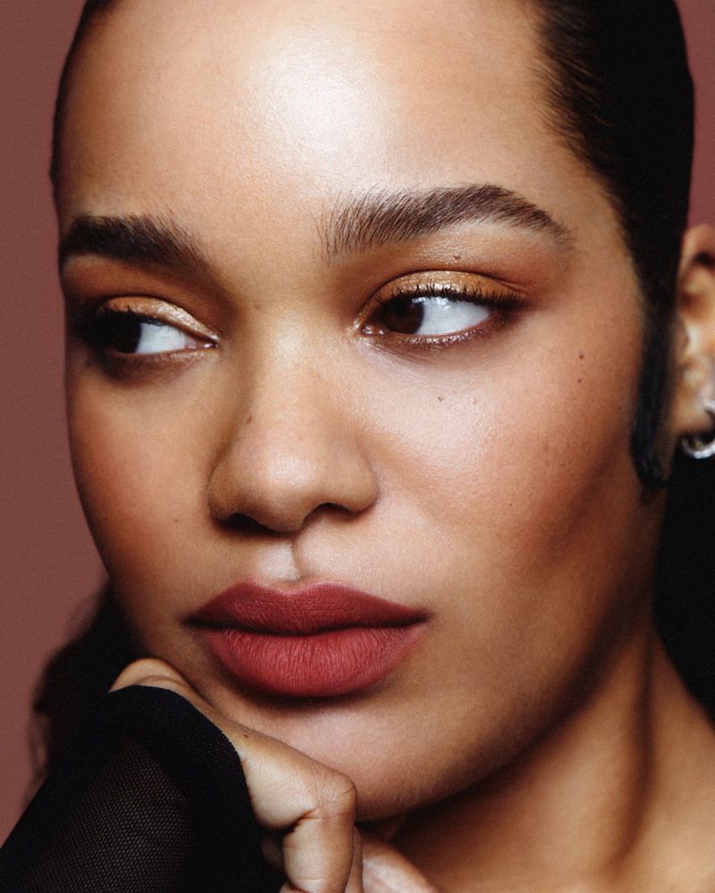 Close-up portrait of a woman with brown lipstick and gold eye shadow, resting her chin on her hand, with a diffused reddish-brown background.