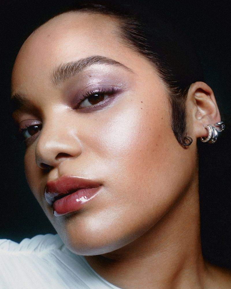 Close-up beauty portrait of model Ariish Wol wearing glossy makeup and silver ear cuffs against a dark background.