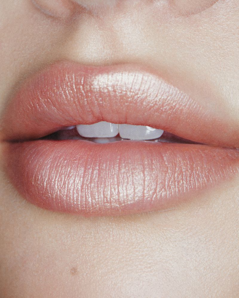 Extreme close-up of glossy pink shimmery lips slightly parted to reveal front teeth against smooth skin.