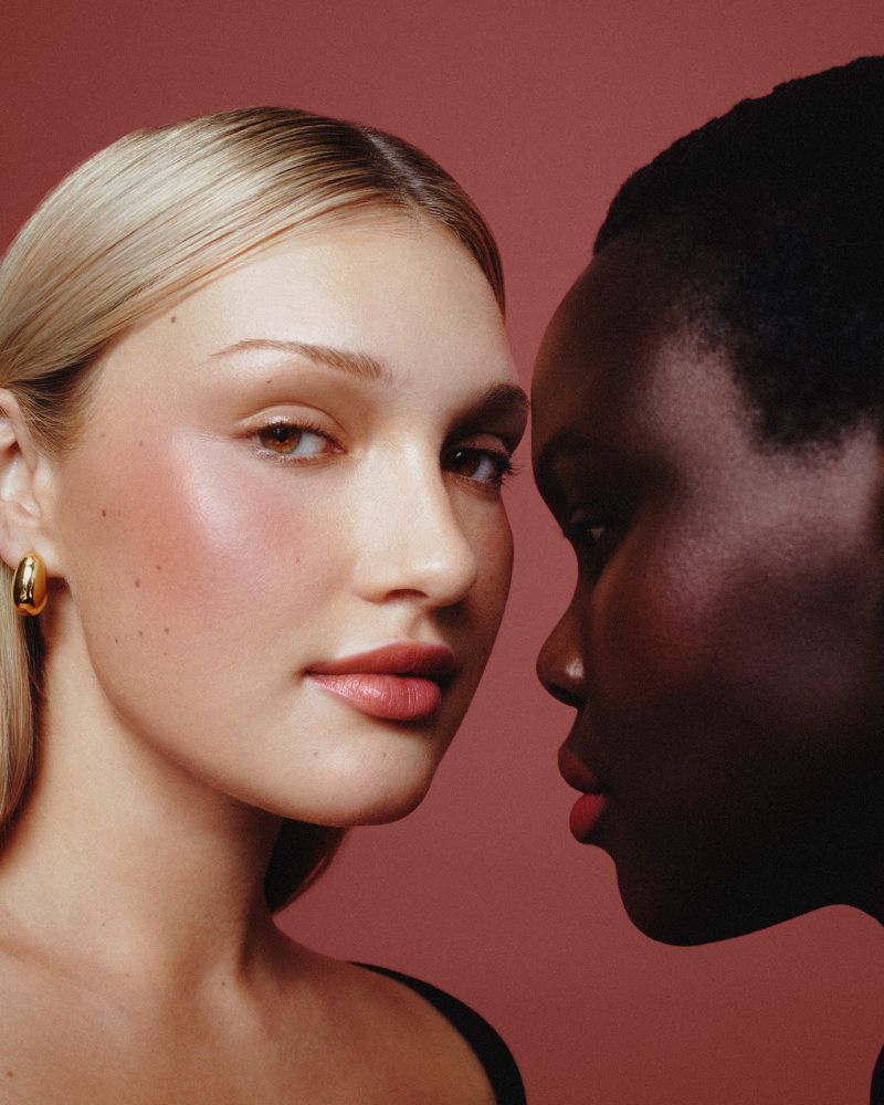Close-up shot of two women with different skin tones posing against a solid pink background in a beauty or fashion context.