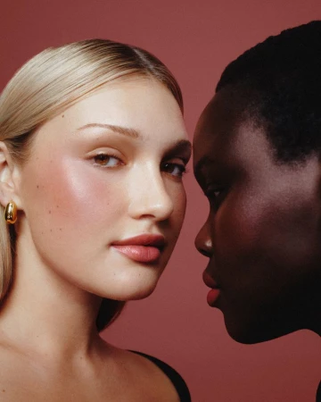 Close-up beauty portrait of two female models with glowing makeup and rosy blush against a dusty pink studio backdrop.