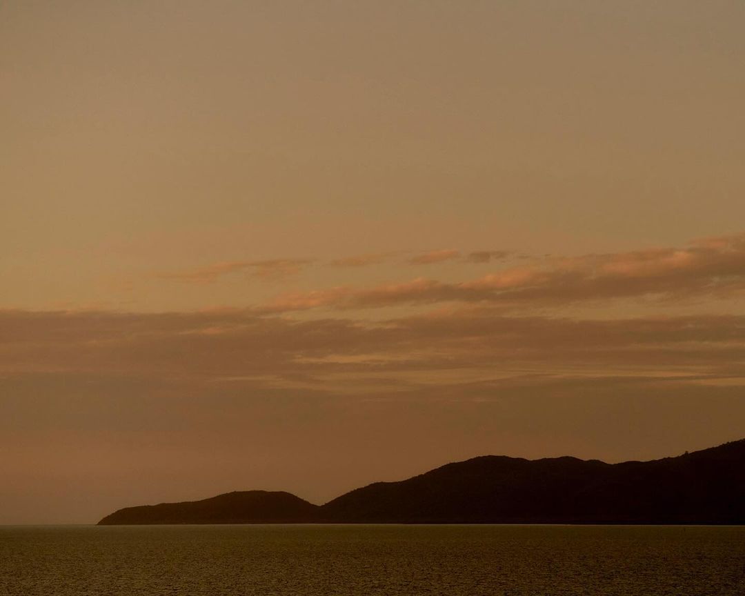 Seascape photo of still water and shadowed land under an orange sky at sunset. Hazy clouds are overhead in the evening sky.