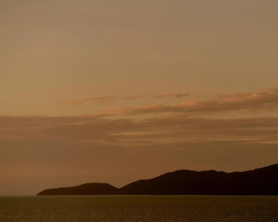 Seascape photo of still water and shadowed land under an orange sky at sunset. Hazy clouds are overhead in the evening sky.
