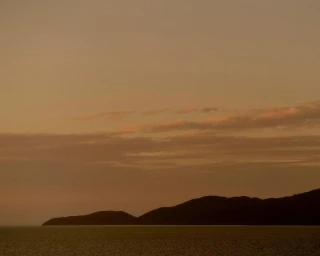 Dark silhouette of rolling hills meeting a calm sea beneath a warm sunset sky filled with layered clouds.