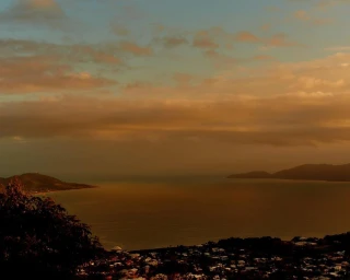 Elevated coastal landscape view overlooking the ocean at sunset with scattered clouds and distant headlands under warm golden light.