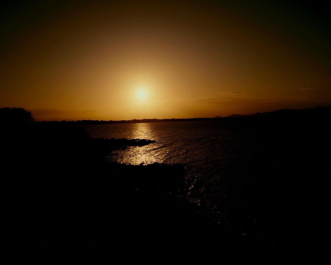Sun setting over a dark body of water with golden sunlight reflecting on the surface and silhouetted land on the horizon.