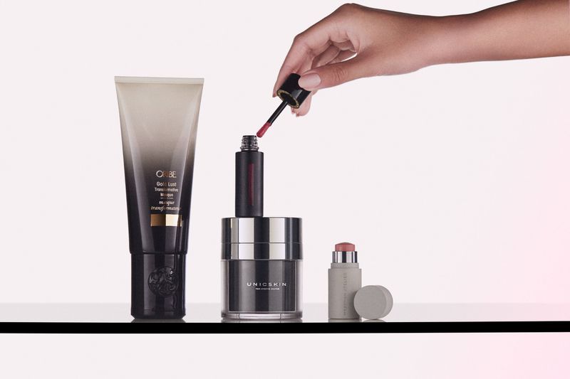 Still life shot of cosmetic products on a glass surface, including a lip gloss, moisturizer, and hair product, with a hand applying lip gloss.