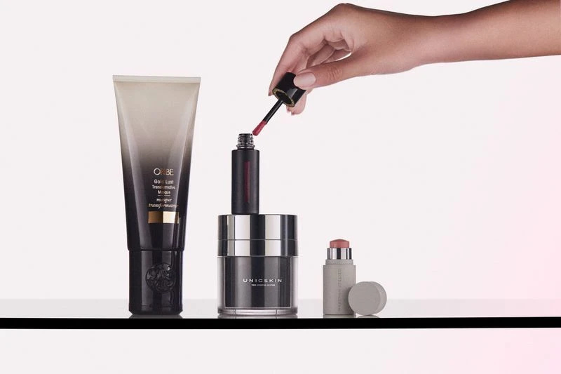 Still life shot of cosmetic products on a glass surface, including a lip gloss, moisturizer, and hair product, with a hand applying lip gloss.