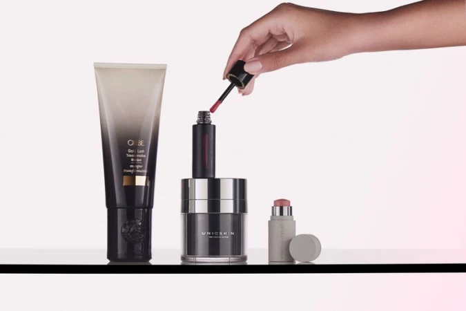 Still life shot of cosmetic products on a glass surface, including a lip gloss, moisturizer, and hair product, with a hand applying lip gloss.