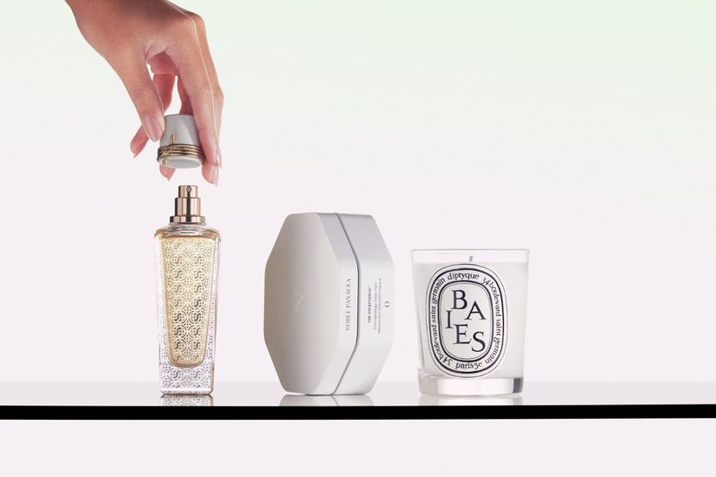 Perfume bottle and candle sit on a reflective surface, with an adult hand placing the cap on the perfume bottle in a photo studio setting.