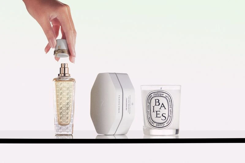 Perfume bottle and candle sit on a reflective surface, with an adult hand placing the cap on the perfume bottle in a photo studio setting.