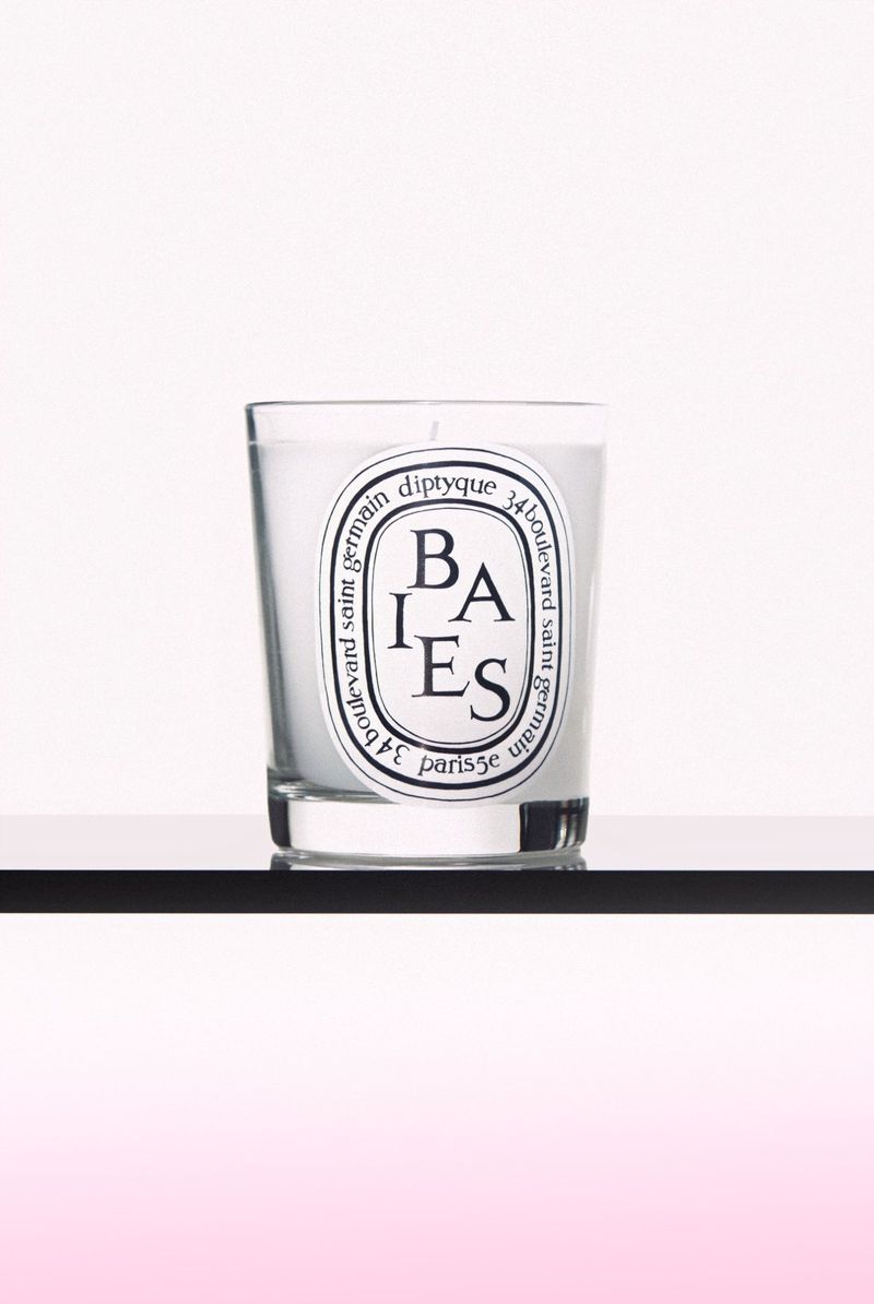Diptyque Baies candle in glass holder with white wax and black and white label on black shelf against a white and pink background.