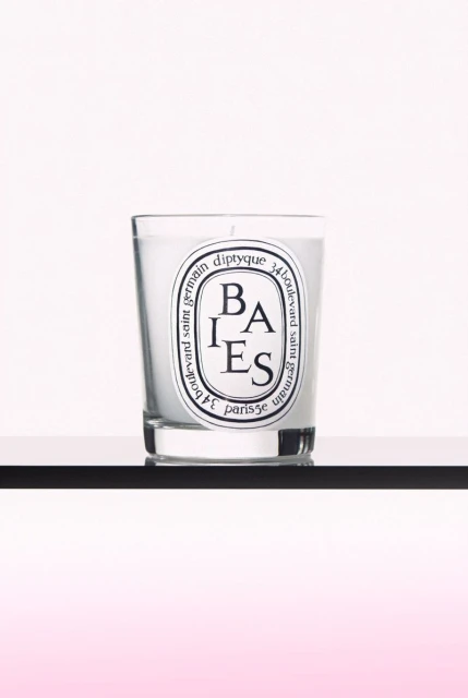 Diptyque Baies candle in glass holder with white wax and black and white label on black shelf against a white and pink background.