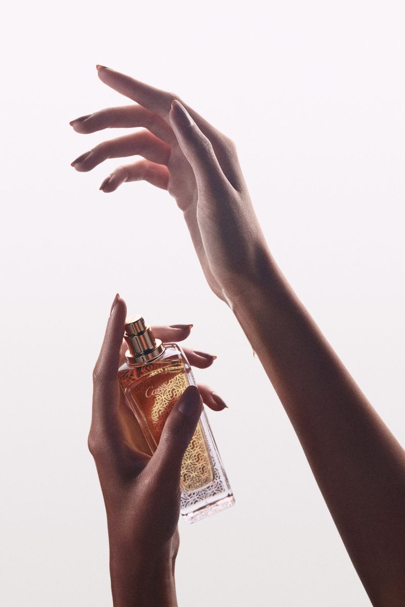Perfume bottle held in hand against a bright white background with gold accents, neutral nail polish, and soft lighting.