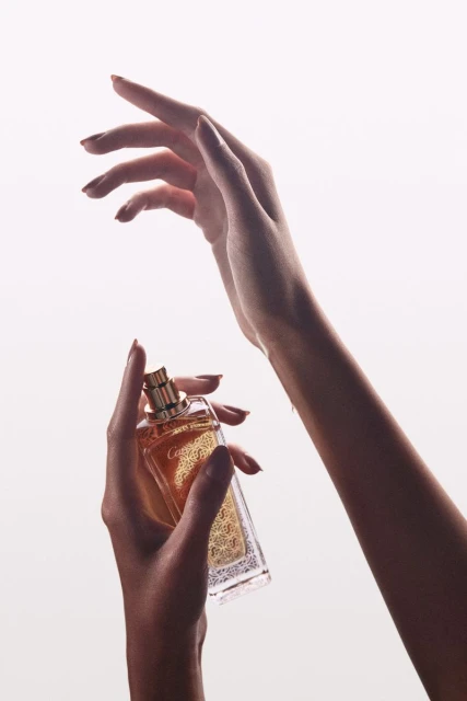 Perfume bottle held in hand against a bright white background with gold accents, neutral nail polish, and soft lighting.