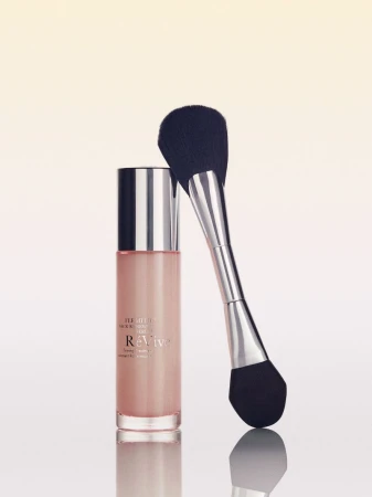 Dual-ended makeup brush leaning against a pink liquid skincare bottle with a silver cap on a reflective surface.