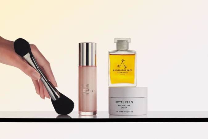 Hand holding a double-ended makeup brush beside luxury skincare serum, oil, and cream jars on a reflective surface.