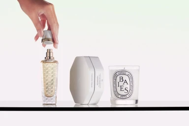 Hand lifting the cap off an ornate perfume bottle beside a white skincare jar and a scented candle on a white surface.