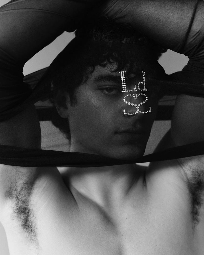 Monochrome portrait of a shirtless man with curly hair, adorned with rhinestone lettering, posing against a plain, light-colored backdrop.