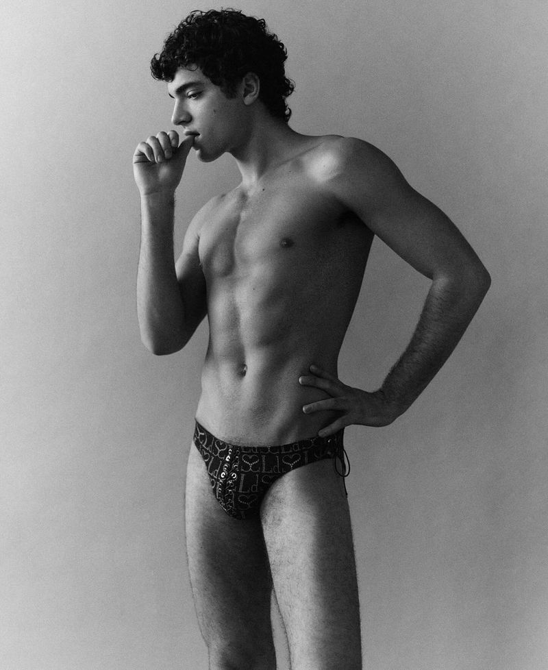 Monochrome portrait of a muscular, bare-chested young man with curly hair, hand to his mouth, wearing patterned swimwear bottoms against a plain wall.
