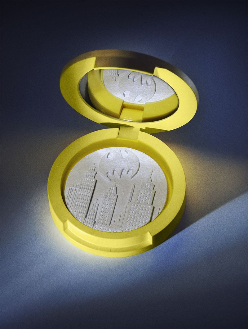 Yellow compact makeup case with Batman logo and city skyline design, spotlighted against a blue background in a studio shot.