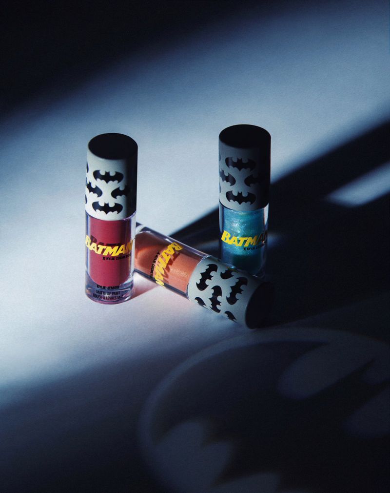 Three tubes of Batman themed lip gloss stand against a gradient white and blue background with the logo of Batman in the corner.