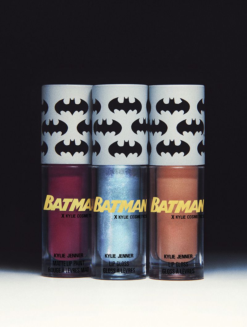 Three Kylie Cosmetics lip glosses in red, blue, and orange with Batman-themed caps, set against a gradient black and white background.