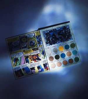 Kylie Cosmetics Batman collaboration eyeshadow palette styled as an open comic book with fifteen colorful shades under spotlight.