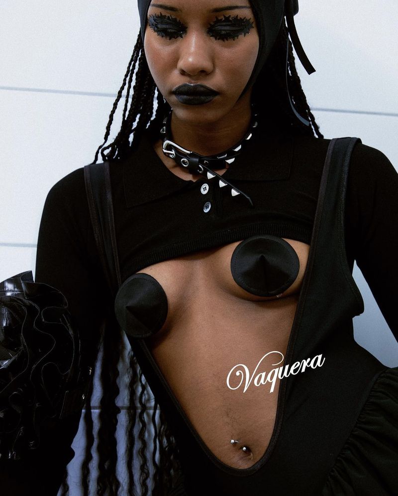 Fashion editorial shot of an adult female model in black avant-garde dress with cone bra details and exaggerated makeup, posing indoors.