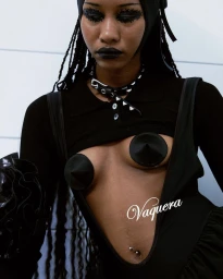 Model wearing dark clothing, studded collar and head wrap with dramatic black makeup against a plain wall