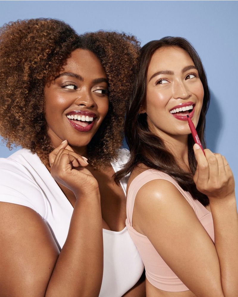 Two women, one African American with curly hair and one Asian applying pink lipstick, smile against a blue background in a beauty shot.