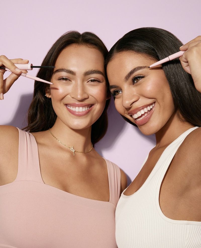 Two smiling young women posing indoors with beauty products for a cosmetics advertisement against a solid purple background.