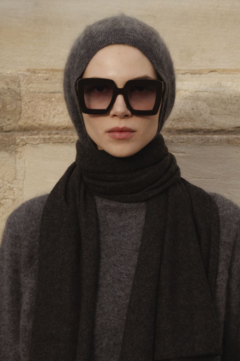 A woman wearing large square sunglasses, a gray knit hat, and a scarf poses against a textured wall in an eye-level portrait.