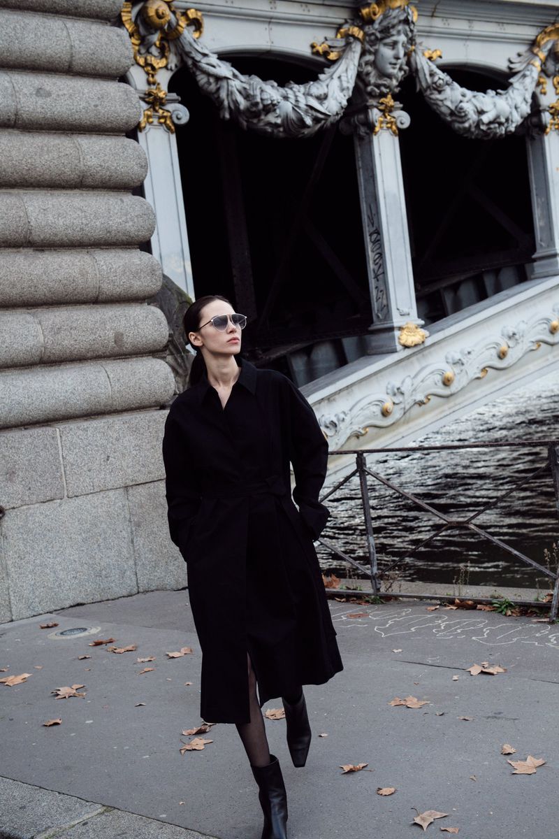 Model wearing dark sunglasses and a black coat walking near an ornate bridge and stone walkway.