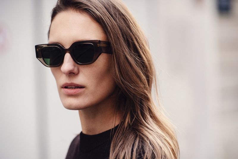 A close-up of a woman with long brown hair wearing sunglasses in an outdoor setting with a blurred background.