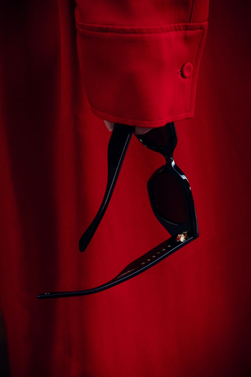 Close-up of a hand holding black acetate sunglasses against a rich red fabric sleeve.