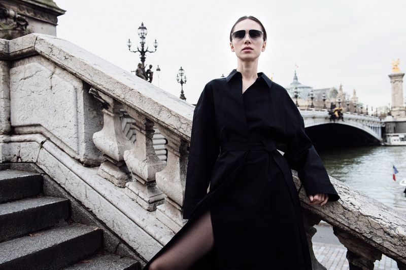 Fashionable woman in sunglasses and black coat stands on a Parisian bridge with stone railings, river, and architecture in the background.