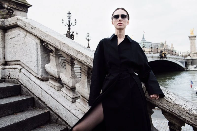 Fashionable woman in sunglasses and black coat stands on a Parisian bridge with stone railings, river, and architecture in the background.