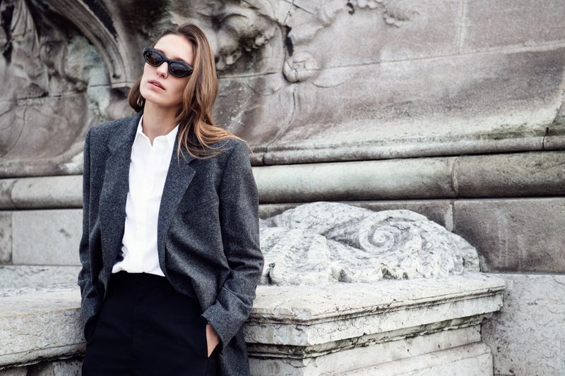 A woman in a grey blazer and black pants poses against a stone wall wearing sunglasses and a white top in an outdoor environment.
