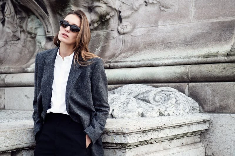 A woman in a grey blazer and black pants poses against a stone wall wearing sunglasses and a white top in an outdoor environment.