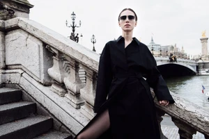 Model in sunglasses wearing a black coat standing on stone stairs by a bridge