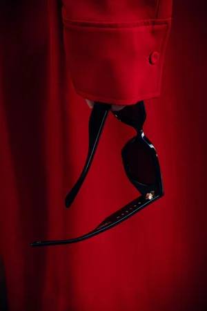 Close-up of a hand holding black acetate sunglasses against a rich red fabric sleeve.